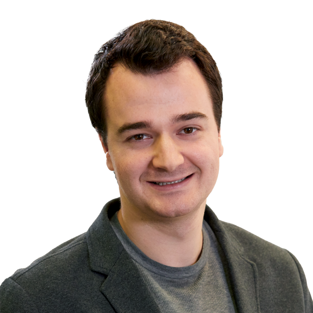 Headshot of Justin, a white man with dark brown hair. Justin is wearing a deep green tweed jacket over a heather gray ribbed shirt, and is smiling towards the camera.